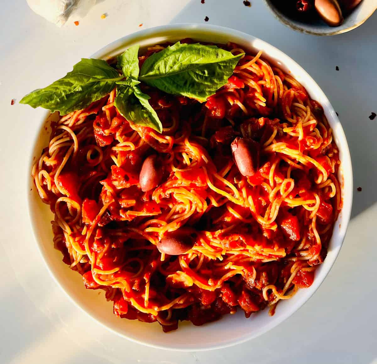 Large white bowl filled with angel hair pasta and vegan puttanesca sauce with basil leaves. Bowl is on a white surface.