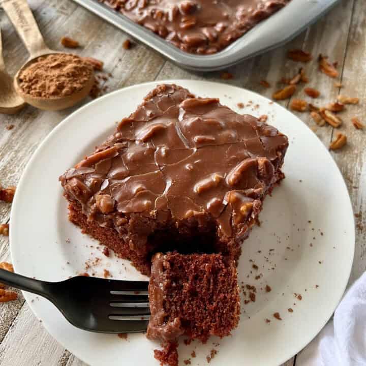There is a white plate with a piece of chocolate cake with chocolate frosting. There is a black fork with a piece of cake on it resting on the plate. There is a measuring spoon with cocoa powder in it and pieces of pecans lying on the surface. There is the corner of a baking pan with cake in it, in the background. Items are on a wooded surface.