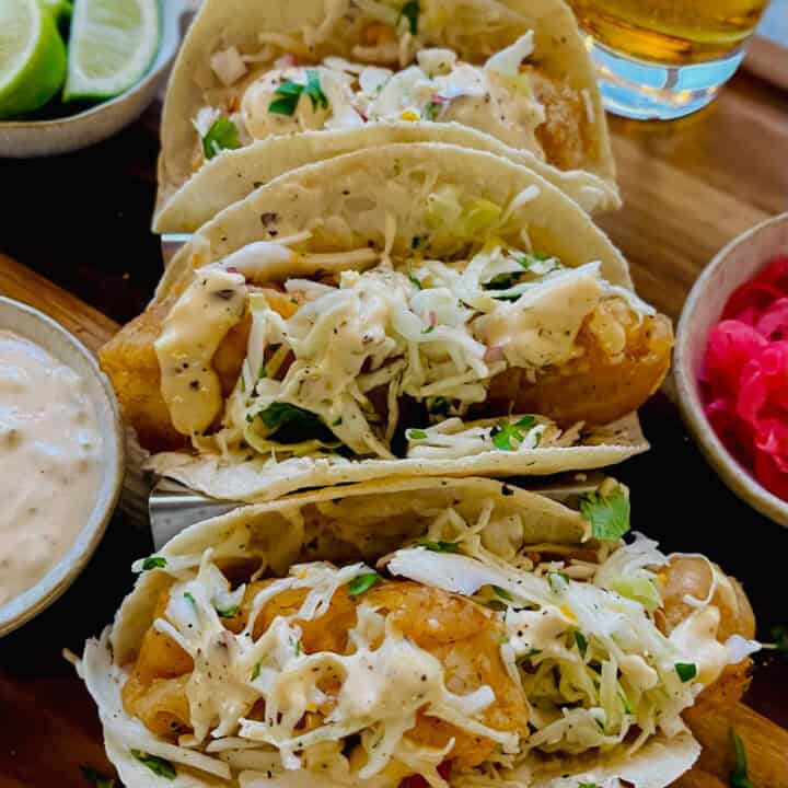 Three fried fish tacos with cabbage slaw and a cream sauce in a taco stand. There is a bowl of pickled red onions, a bowl of lime wedges, a bowl of cream sauce, and a glass of beer. Items are on a wood surface.