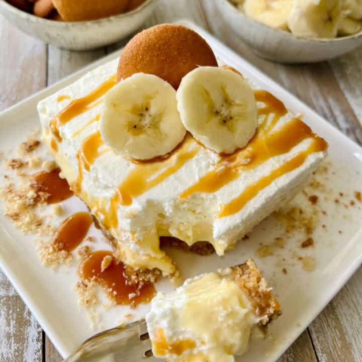 White square plate with a piece of banana cheesecake on it. There is a fork with a bite of cheesecake resting on the plate. There is caramel sauce drizzled over it and slices of bananas and a cookie on top. There is a small bowl of cookies and another with sliced bananas. Items are on a wood surface.