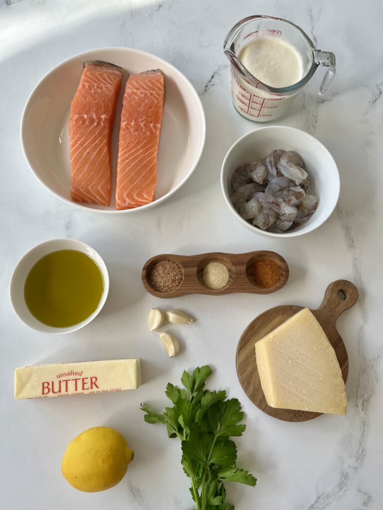 Dishes with ingredient for Cajun Salmon and Shrimp: Large white bowl with two filets of raw salmon, a glass measuring cup with heavy cream, a white bowl with raw shrimp, a white bowl with oil, a wooden dish with dry seasonings in it, a small round wooden board with a block of parmesan cheese, a stick of butter, a whole lemon and sprigs of fresh parsley. Items are on a white surface.