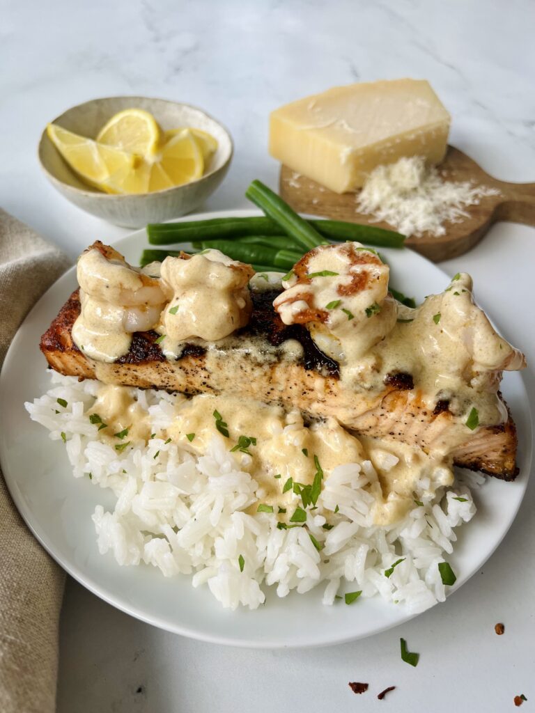 White plate with a bed of white rice. There is a salmon filet topped with shrimp on top of the rice. There is sauce drizzled over the seafood and rice. There are a few green beans on the plate. Chopped parsley sprinkled over the dish. There is a light brown cloth napkin next to the plate. In the background there is a small bowl with lemon wedges and a small round wooden board with parmesan cheese on it. Items are on a white surface.