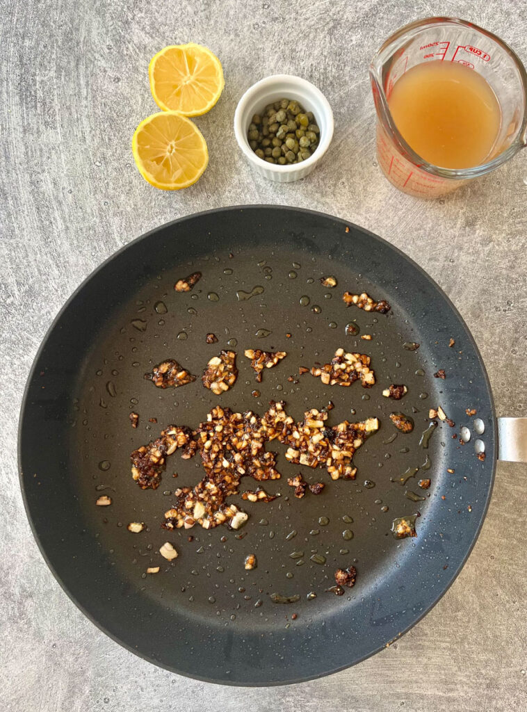 Large gray skillet with cooked garlic and shallots. There is a cut lemon, small dish of capers, and a glass measuring cup with brown broth in it. Items are on a gray surface.