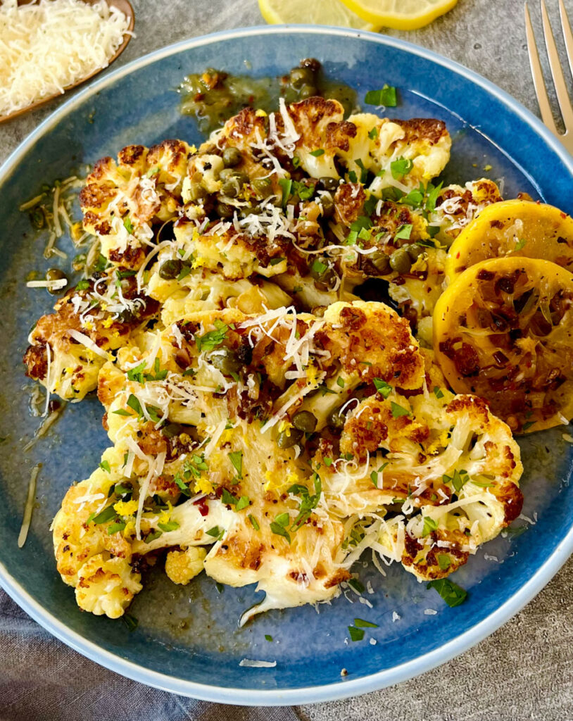 Light blue plate with slices of cauliflower piccata and lemon slices. There is grated parmesan cheese and chopped parsley sprinkled over the dish. Plate is on a gray surface.