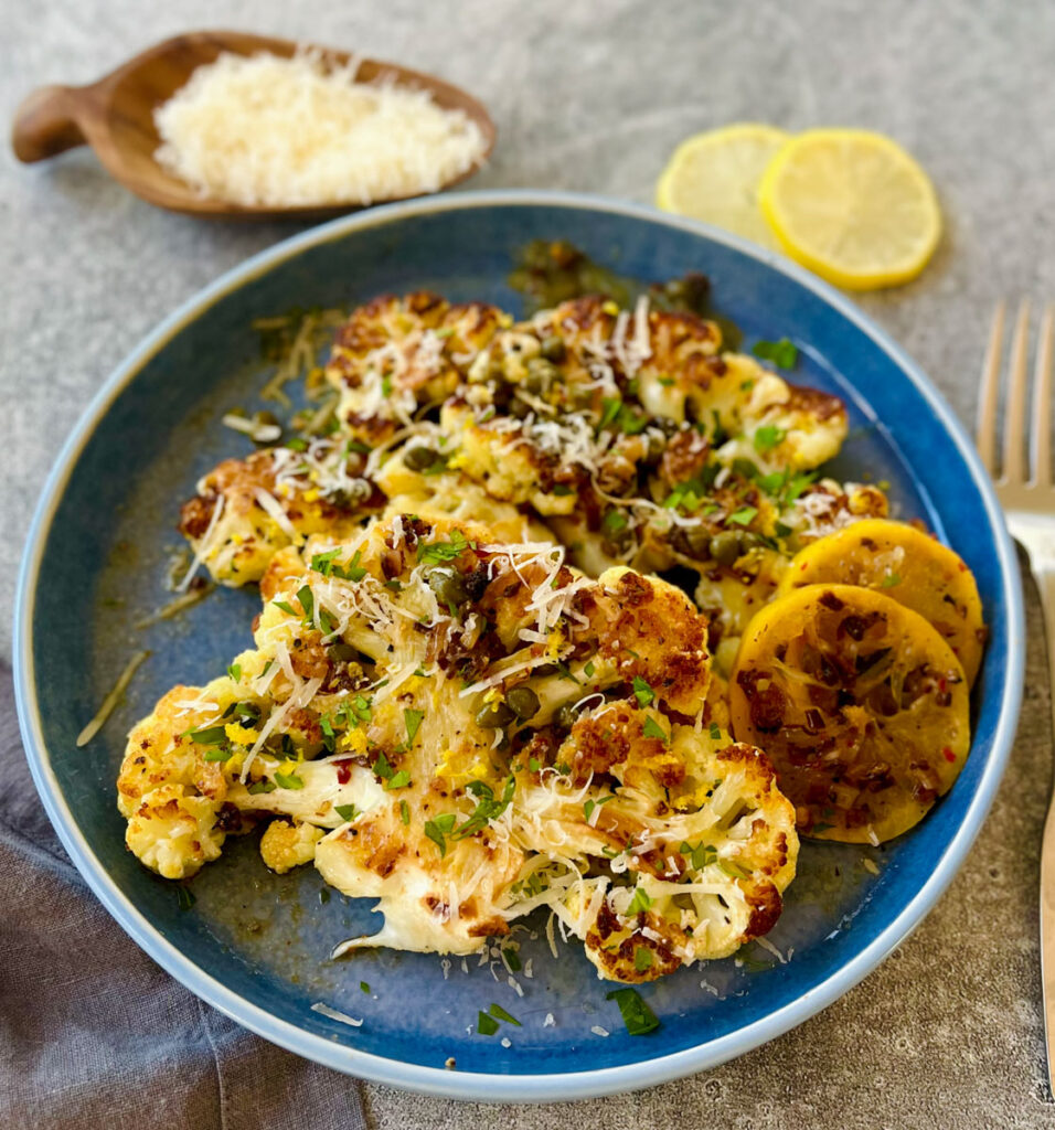 Light blue plate with slices of cauliflower piccata and lemon slices. There is grated parmesan cheese and chopped parsley sprinkled over the dish. There is a gray cloth napkin, a fork, lemon slices and a wooden bowl with grated parmesan cheese in it. Items are on a gray surface.