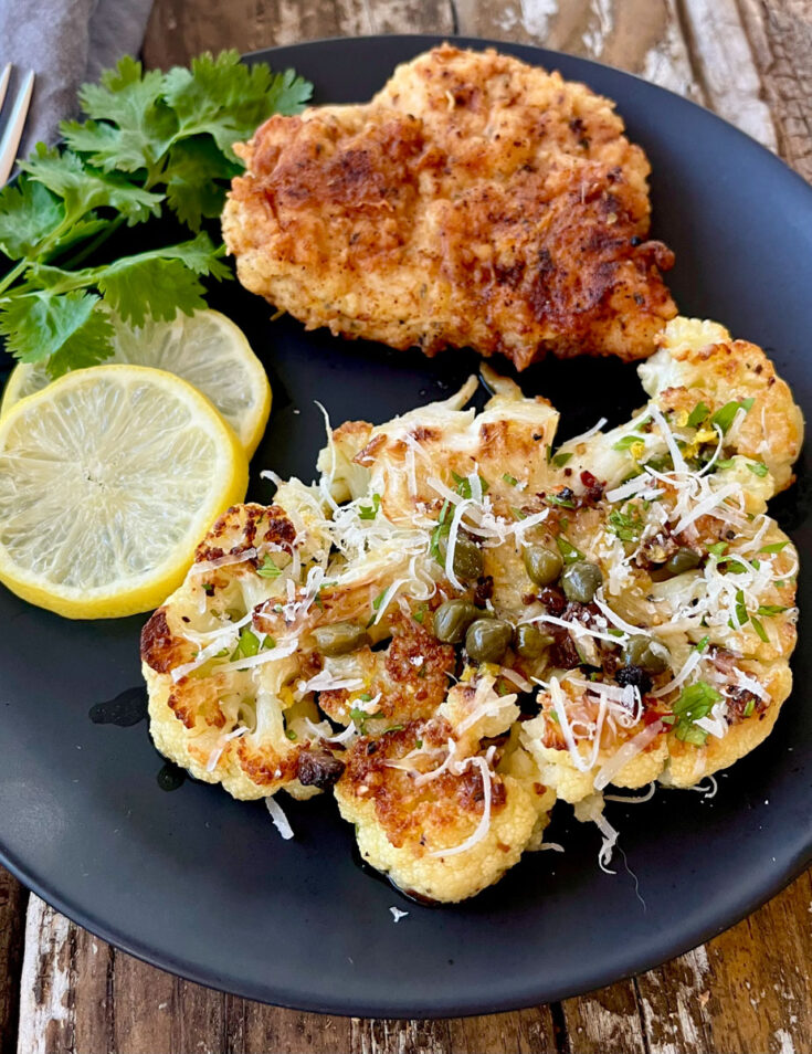 Black plate with a piece of cauliflower steak piccata, breaded chicken breasts. lemon slices, and parsley on it. Plate is on a wood surface.
