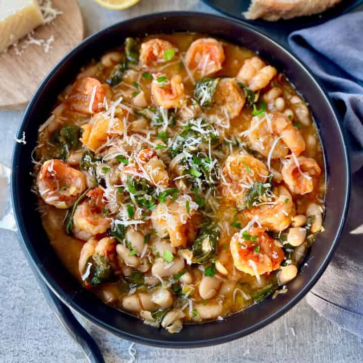 Large black bowl with shrimp and bean stew in it. There is a spoon laying next to the bowl. There is a block of cheese, part of a lemon, a piece of bread on a black plate, and a gray cloth napkin. Items are on a gray surface.