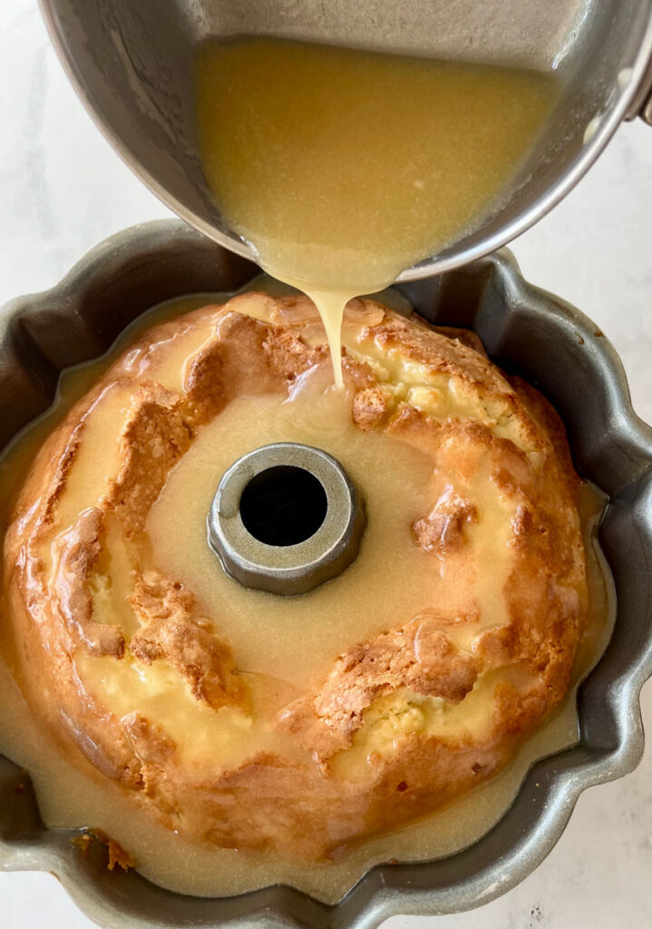 Baked bundt cake in a pan. There is a saucepan with a mixture being poured ver the cake. Pan is on a white surface.