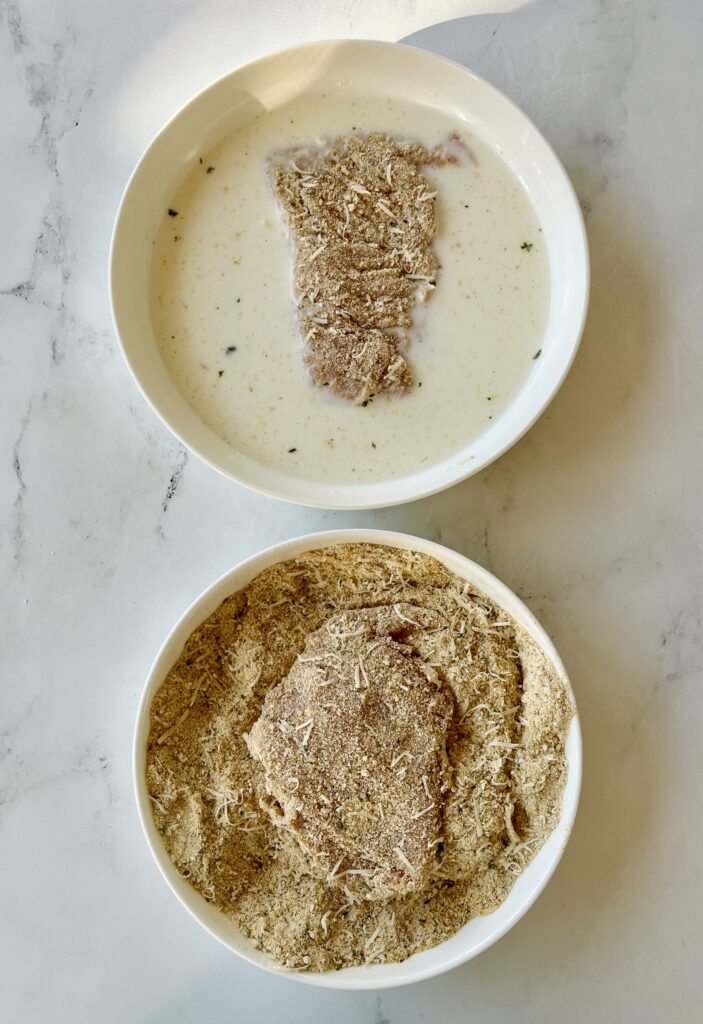 Two large white dishes. One dish has milk in it with a piece of raw breaded chicken. The other dish has a dry breading mixture with a piece of raw chicken in it. Dishes are on a whte surface.