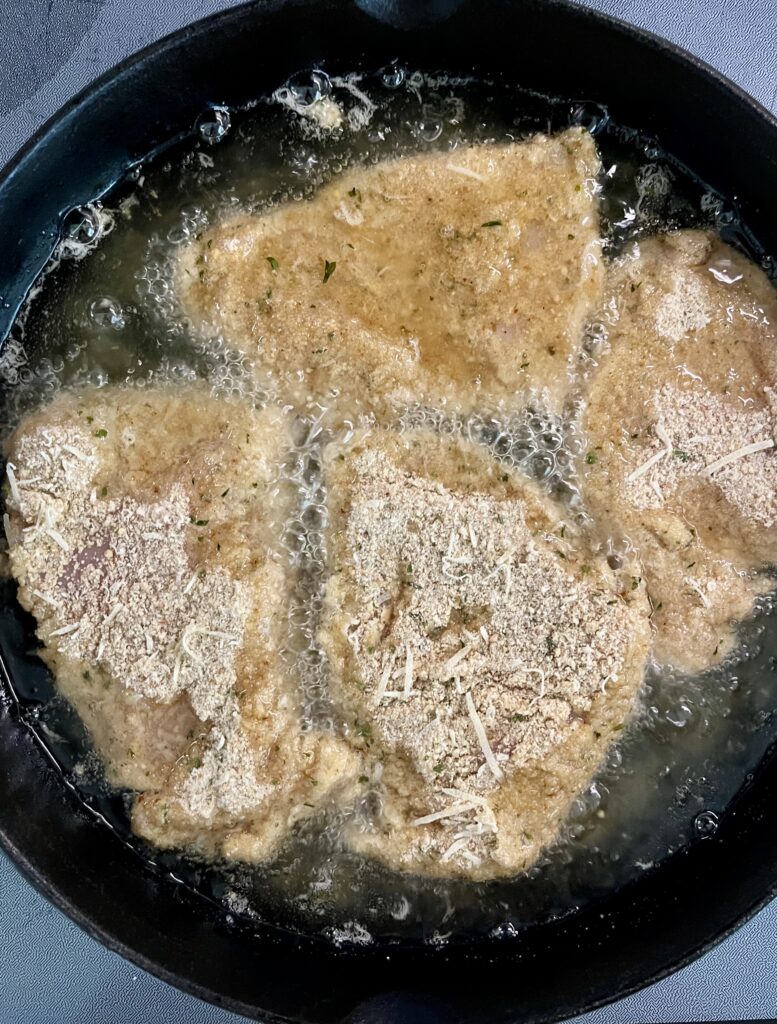 Black iron skillet with four pieces of chicken cooking in oil. Pan is on a gray surface.
