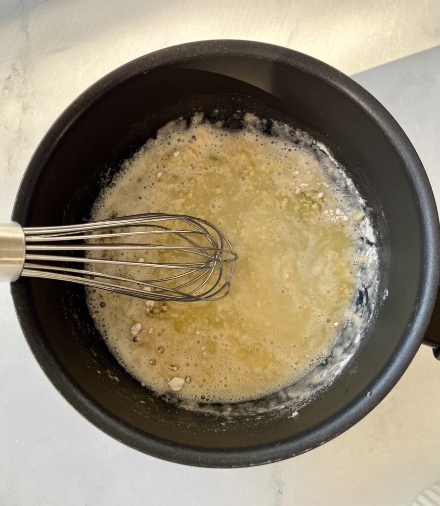 Dark saucepan with melted butter, flour, and minced garlic. There is a whisk resting in the pan. Pan is on a white surface.