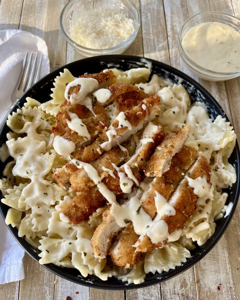 Black bowl with bow tie pasta, pieces of breaded chicken, and white cream sauce. In the background there is a small glass dish with grated white cheese and another with white cream sauce in it. There is a fork and a white cloth napkin. Items are on a wood surface.