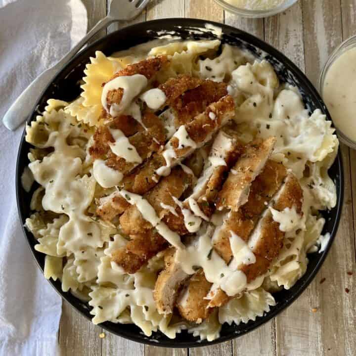 Black bowl with bow tie pasta, pieces of breaded chicken, and white cream sauce. In the background there is a small glass dish with grated white cheese and another with white cream sauce in it. There is a fork and a white cloth napkin. Items are on a wood surface.