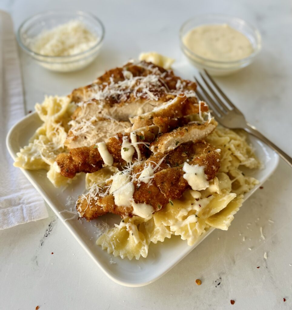 White square plate with bow tie pasta, pieces of breaded chicken, and white cream sauce. There is a fork resting on the plate. In the background there is a small glass dish with grated white cheese and another with white cream sauce in it. There is a white cloth napkin. Items are on a white surface.