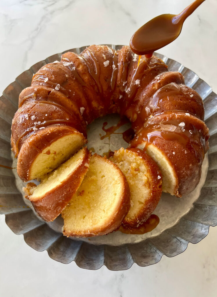 Round bundt cake partially sliced on a round tin dish. There is a spoon with caramel sauce drizzling over the cake. Dish is on a white surface.