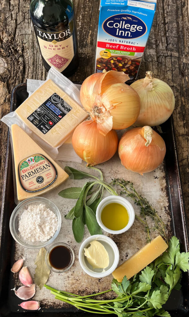 Baking sheet with various ingredients on it: Onions, blocks of cheese, sprigs of sage, parsley, and thyme, garlic cloves, a bay leaf, small dishes with butter, oil, flour in them. There is a bottle of sherry and a box of beef broth. Items are on a wood surface.