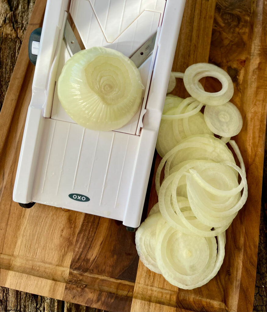 There is a slicing device (mandolin) with an onion on it. There are thin slices of onion. Items are on a wooden board.
