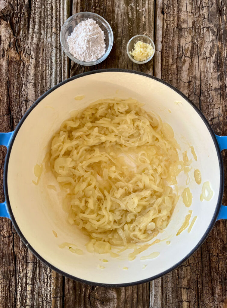 Large pot with cooked onions in it. There is a small dish with flour in it and another with minced garlic in it. Items are on a wood surface.