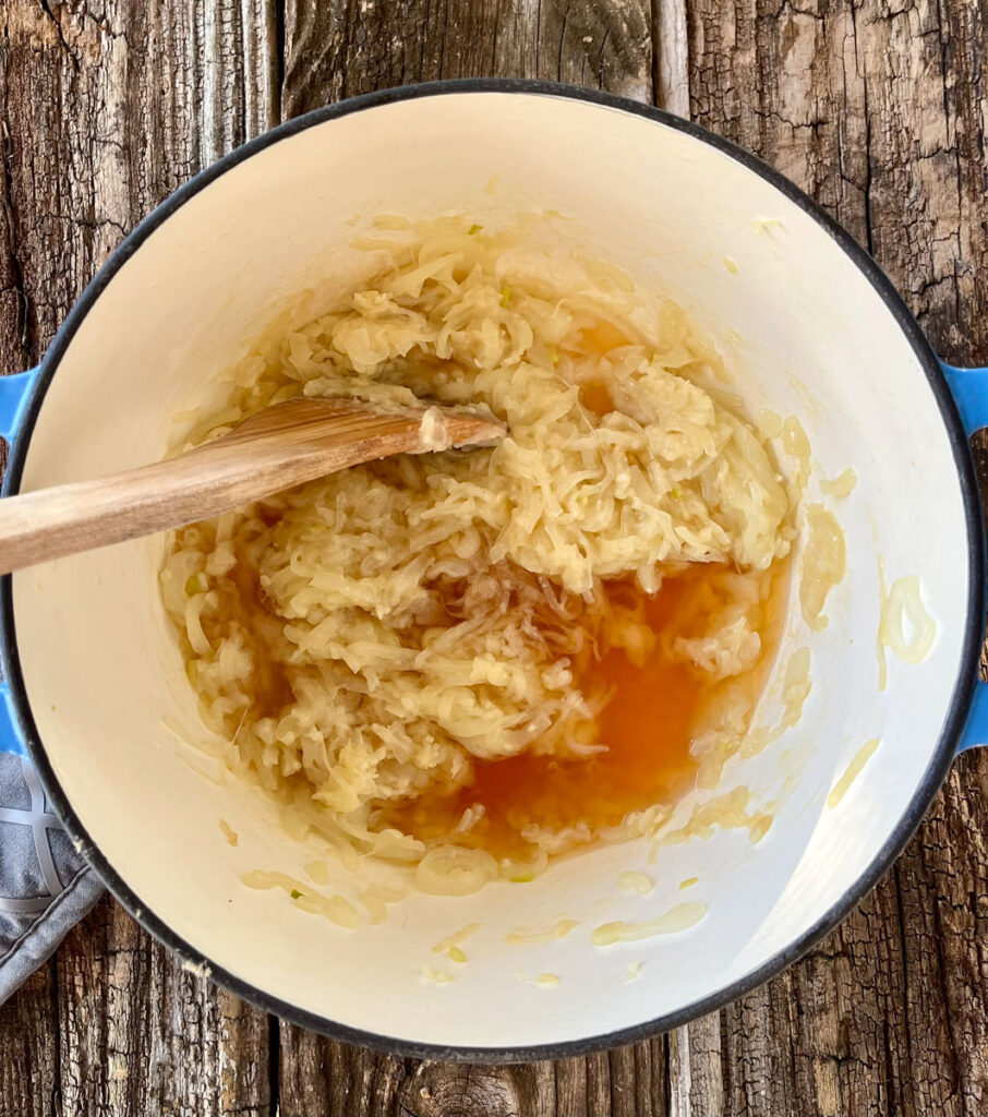 Large pot with an onion mixture and brown liquid in it. There is a wooden spoon resting in the pot. Pot is on a wood surface.