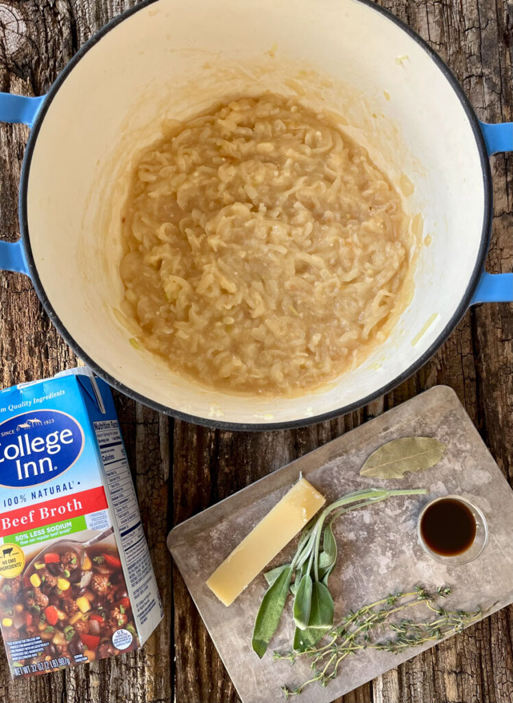Large pot with a cooked onion mixture in it. There is a box of beef broth and a small metal sheet pan with fresh herbs and a cheese rind. Items are on a wood surface.
