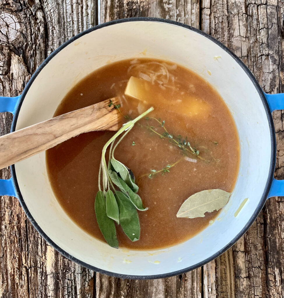 Large pot with a cooked onion, fresh herbs, and beef broth mixture in it. There is a wooden spoon resting in the pot. Pot is on a wood surface.