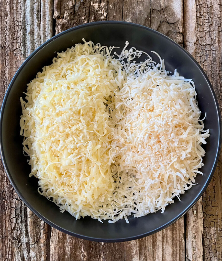 Black shallow bowl with two types of grated cheese. Bowl is on a wood surface.