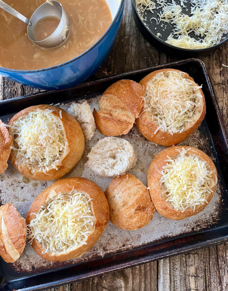 There is a baking sheet with hollowed out bread rounds that have onion soup and grated cheese in them. There is a pot of onion soup, and a bowl of grated cheese. Items are on a wood surface.