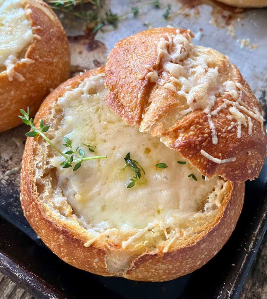 Hollowed out bread round with onion soup topped with melted cheese and fresh herbs. The top of the bread round is leaning against the side. Bread round is on a baking sheet.
