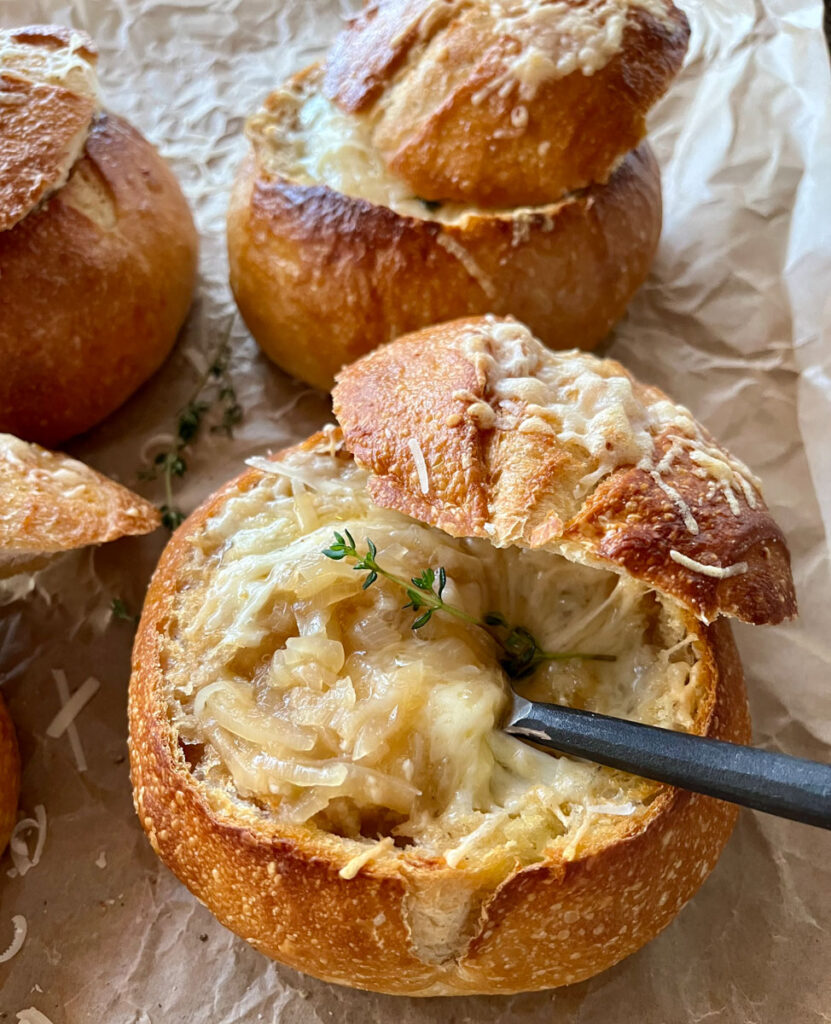 Hollowed out bread rounds with onion soup topped with melted cheese and fresh herbs. The tops of the bread round are leaning against the sides of the rounds. There is a spoon in one of the bread bowls. Bread rounds are on brown paper.