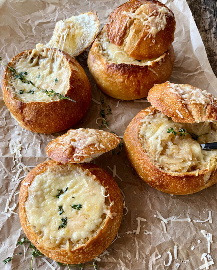 Hollowed out bread rounds with onion soup topped with melted cheese and fresh herbs. The tops of the bread round are leaning against the sides of the rounds. There is a spoon in one of the bread bowls. Bread rounds are on brown paper.