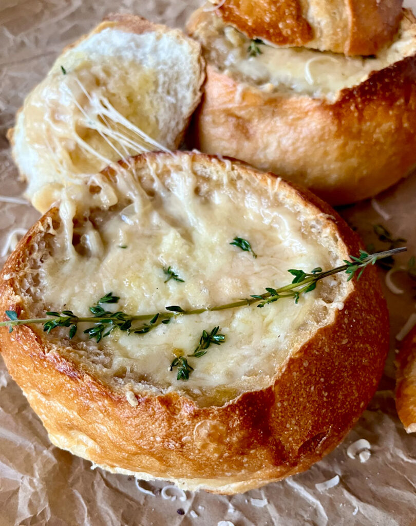 Hollowed out bread round with onion soup topped with melted cheese and fresh herbs. The top of the bread round is leaning against the side. Bread round brown paper.