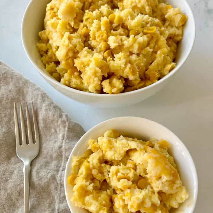 There is a large white bowl with scalloped corn casserole in it. There is a small white bowl with scalloped corn casserole in it and a tan cloth napkin with a fork resting on it. Items are on a white surface.