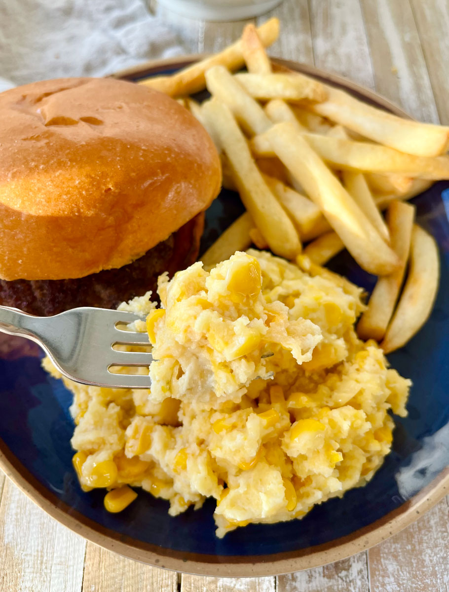 There is a blue plate with scalloped corn casserole, a hamburger, and french fries on it. There is a fork in the scalloped corn. Items are on a wood surface.