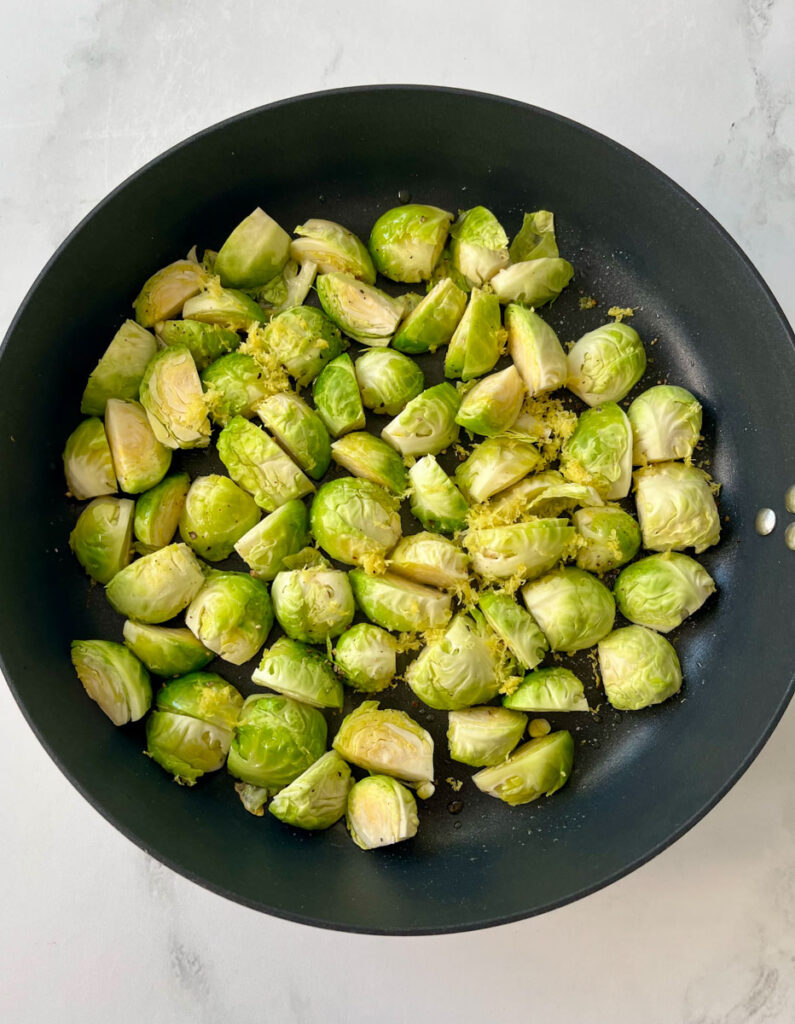 There is a dark skillet with cut up Brussels sprouts olive oil and lemon zest. Skillet is on a white surface.