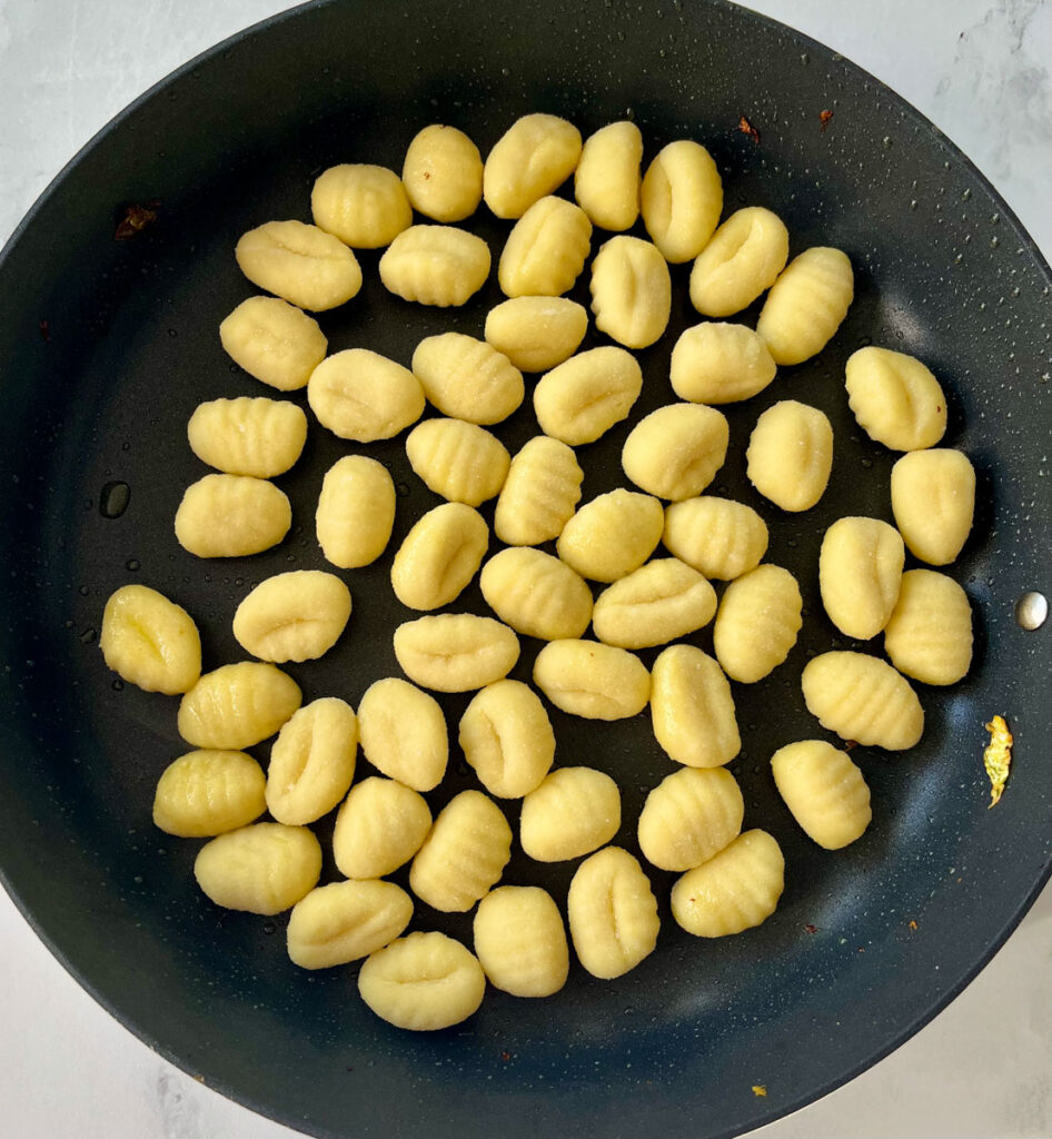 Large skillet with uncooked gnocchi and olive oil in it. Skillet is on a white surface.