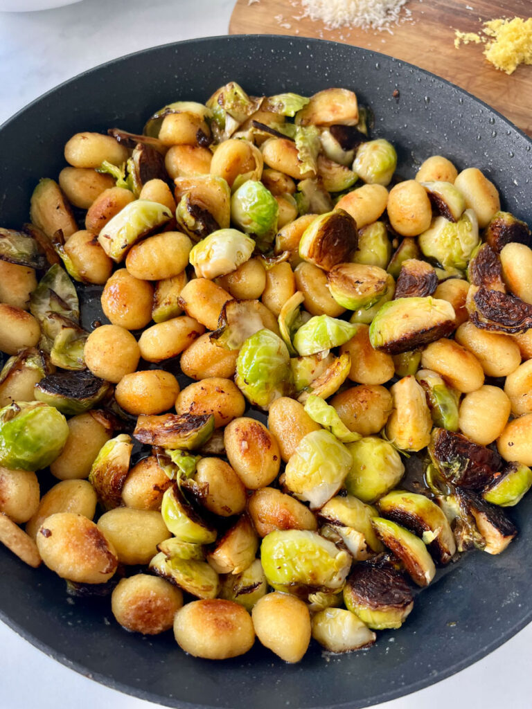 There is a large skillet with cooked gnocchi and Brussels sprouts in it. There is a wooden board. Items are on a white surface.