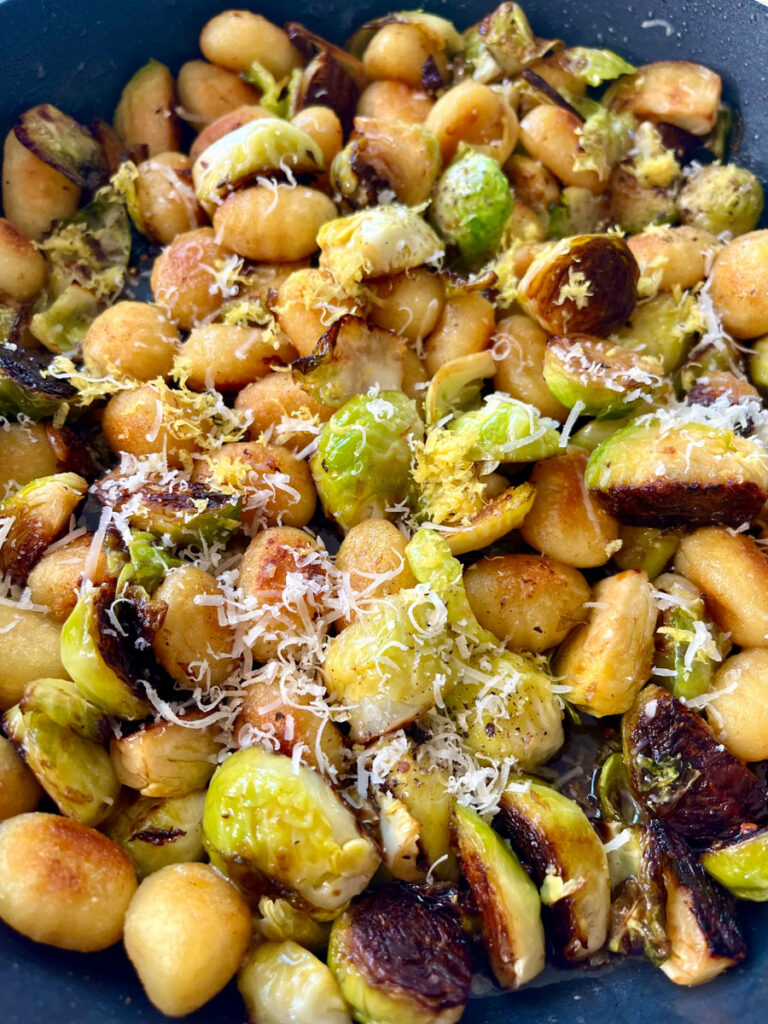There is a large skillet with cooked gnocchi and Brussels sprouts in it. There is grated white cheese sprinkled over the top.
