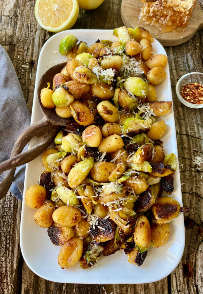 Cooked gnocchi and Brussels sprouts with grated cheese on a white platter. There is a wooden spoon resting on the platter. There is a cut lemon, a gray cloth, a small dish with red pepper flakes. and a small round wood board with a honeycomb on it. items are on a wood surface.