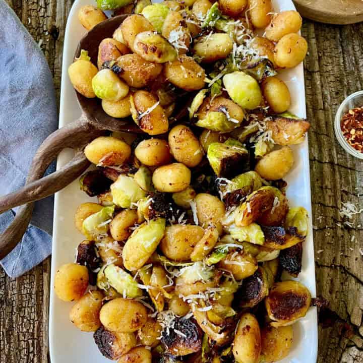 Cooked gnocchi and Brussels sprouts with grated cheese on a white platter. There is a wooden spoon resting on the platter. There is a cut lemon, a gray cloth, a small dish with red pepper flakes. and a small round wood board with a honeycomb on it. items are on a wood surface.
