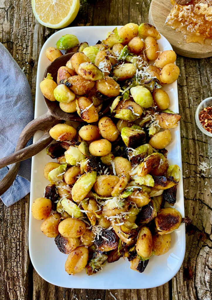 Cooked gnocchi and Brussels sprouts with grated cheese on a white platter. There is a wooden spoon resting on the platter. There is a cut lemon, a gray cloth, a small dish with red pepper flakes. and a small round wood board with a honeycomb on it. items are on a wood surface.