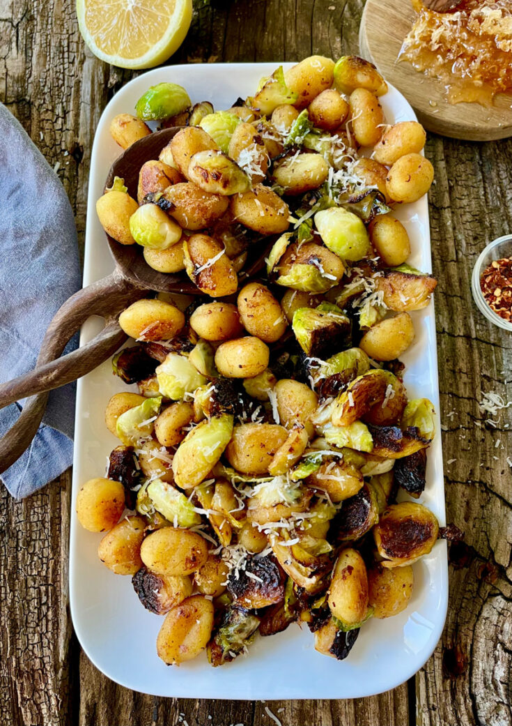 Cooked gnocchi and Brussels sprouts with grated cheese on a white platter. There is a wooden spoon resting on the platter. There is a cut lemon, a gray cloth, a small dish with red pepper flakes. and a small round wood board with a honeycomb on it. items are on a wood surface.