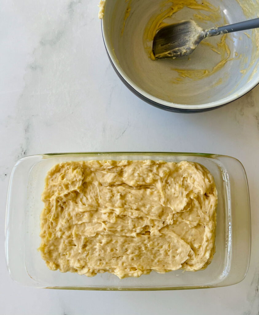There is a clear glass baking pan with a yellow bread batter in it. There is an empty large bowl with a gray rubber spatula resting in it in the background. Items are on a white surface.