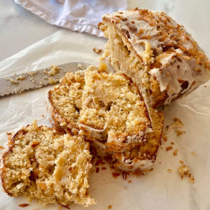 There is a loaf of sliced bread with white frosting and toasted shredded coconut on top. The bread is on white paper. There is a large knife next to the bread. There is a white cloth napkin in the background. Items are on a white surface.