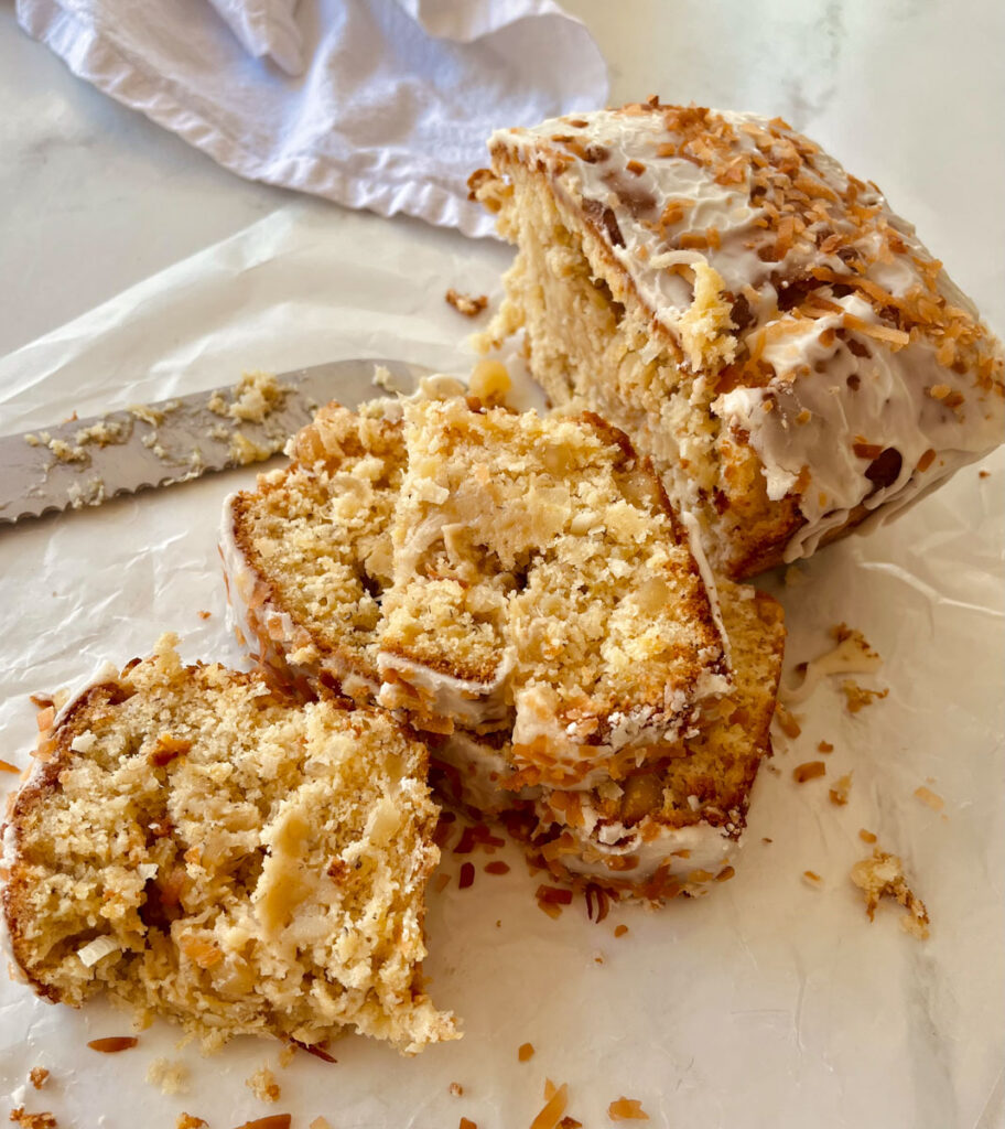 There is a loaf of sliced bread with white frosting and toasted shredded coconut on top. The bread is on white paper. There is a large knife next to the bread. There is a white cloth napkin in the background. Items are on a white surface.