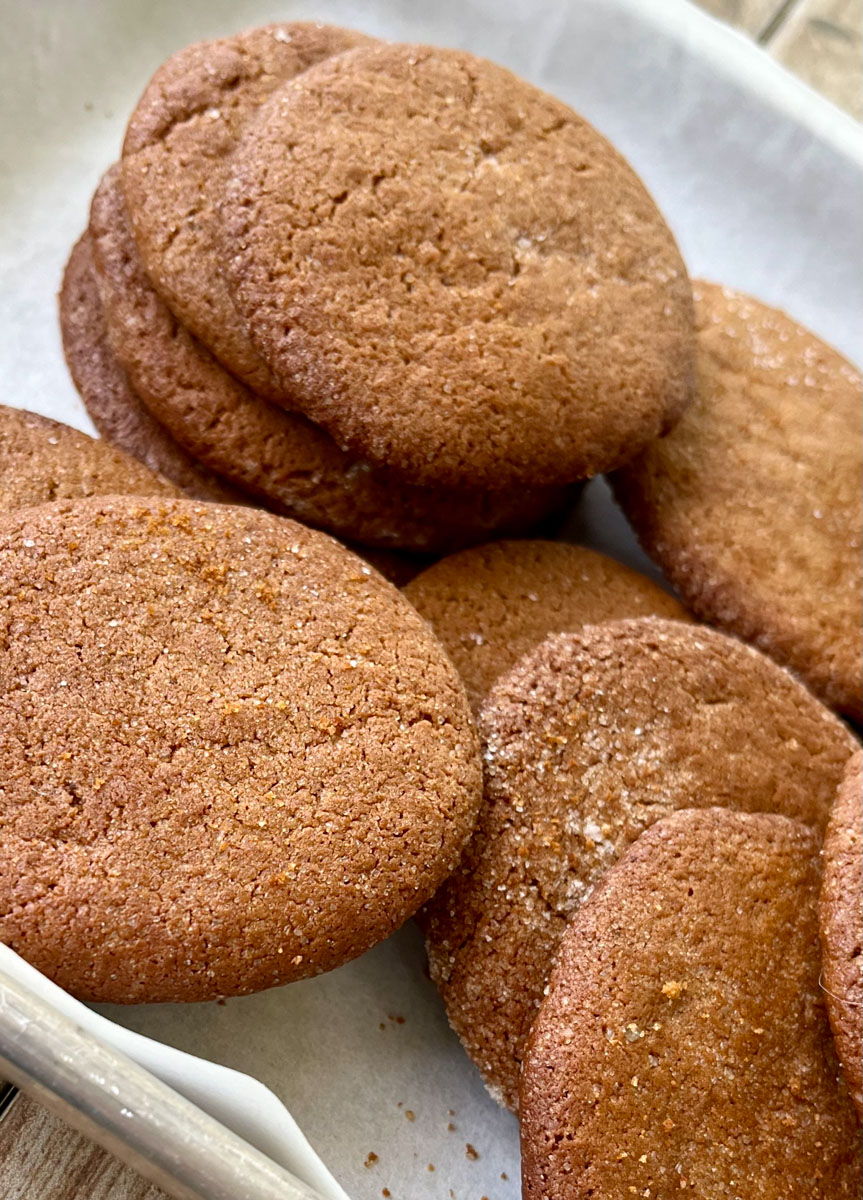 Thin brown cookies on a white paper-lined tray. Tray is on a wood surface.