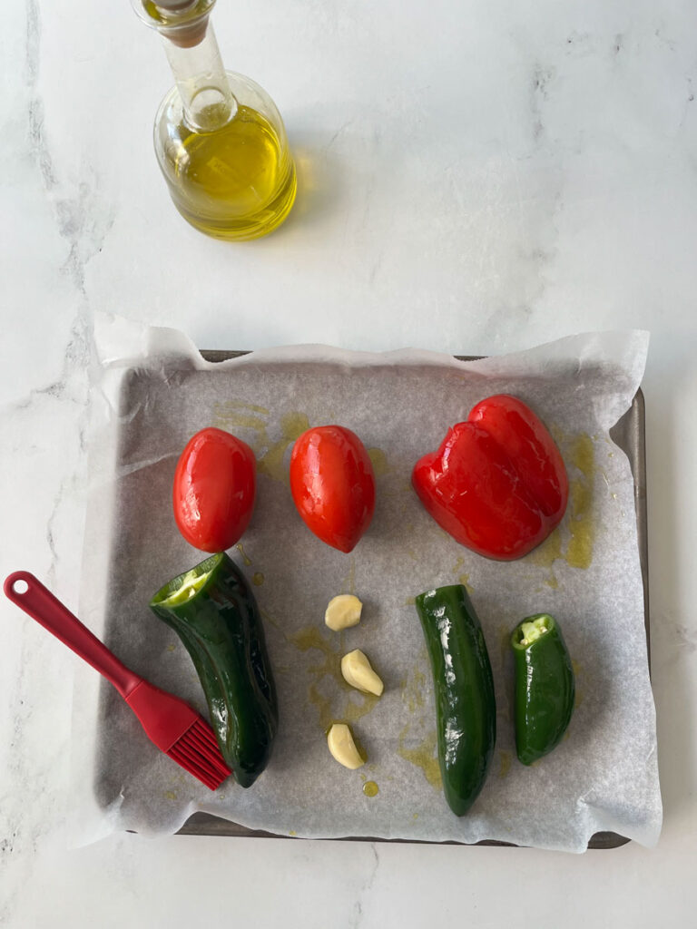 There is a silver baking sheet lined with white paper. There are two tomatoes, a red bell pepper, three long green peppers, three garlic cloves. There is oil drizzled over the items on the pan. There is a red basting brush resting on the pan. In the background there is a clear bottle of cooking oil. Items are on a white surface.