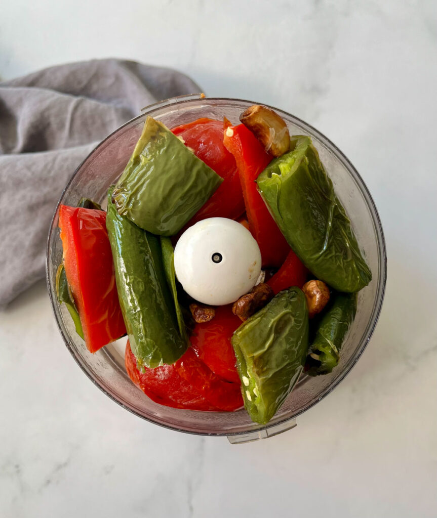 There is a food processor (chopper) bowl with roasted peppers and garlic in it. There is a gray cloth napkin. Items are on a gray surface.