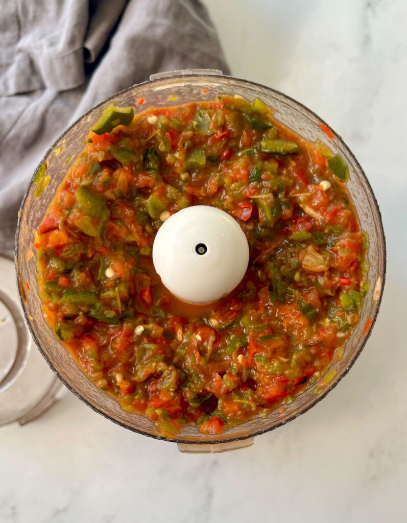 There is a food processor (chopper) bowl with a vegetable and pepper chopped mixture in it. There is a gray cloth napkin. Items are on a gray surface.