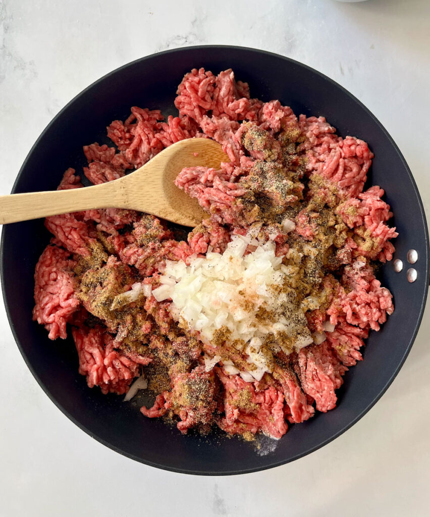 There is a a large dark skillet with ground beef, chopped onions and dry seasonings cooking. There is a wooden spoon resting in the skillet. Skillet is on a white surface.