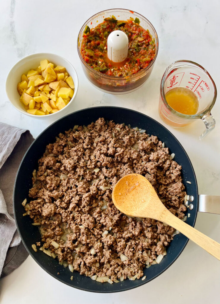 There is a large dark skillet with ground beef, and onions in it. There is a pile of chopped potatoes on the mixture. There is a wooden spoon resting in the pan. There is a gray cloth napkin laying next to the pan. In the background there is a white bowl with chopped potatoes in it, a round bowl of a food processor (chopper) with a pepper and vegetable mixture in it and a glass measuring cup with a brown liquid in ti. Items are on a white surface.